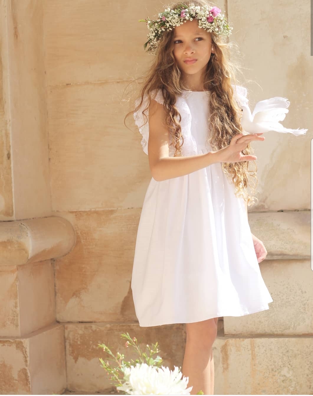 Menina com vestido branco e coroa de flores, em ambiente exterior com parede de pedra clara