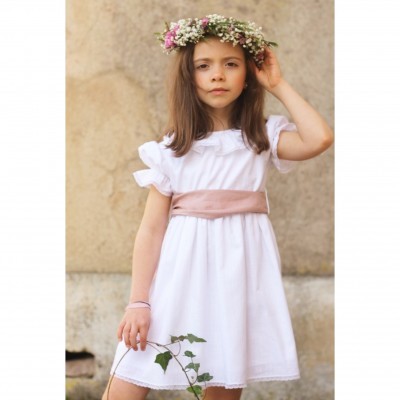 Menina com vestido branco e cinto rosa claro, coroa de flores e ramo verde, em fundo de parede bege