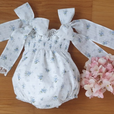 Conjunto de roupa infantil com padrão floral azul sobre fundo branco, mangas com laços e renda, ao lado de flores cor-de-rosa
