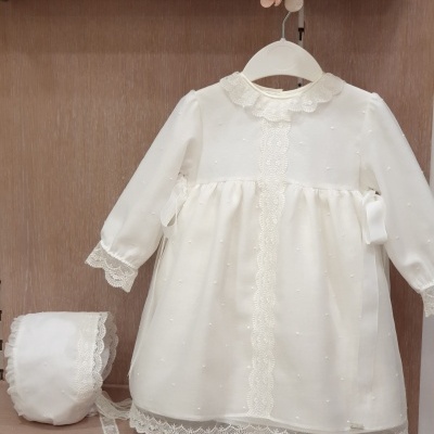 Vestido branco de bebé com renda e gorro branco ao lado, pendurado num cabide