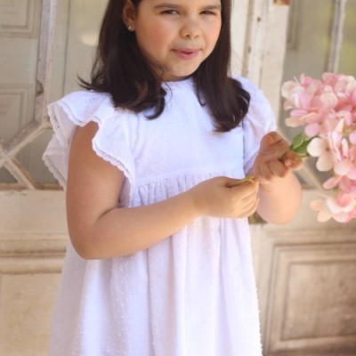 Menina vestindo vestido branco com textura de bolinhas e mangas curtas, segurando flores cor-de-rosa