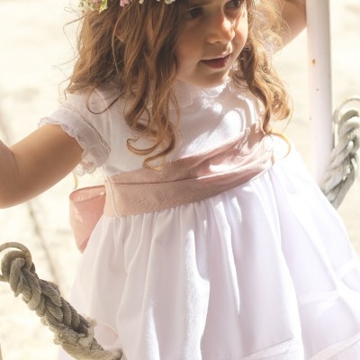 Criança com vestido branco e faixa rosa numa baloiço com coroa de flores.