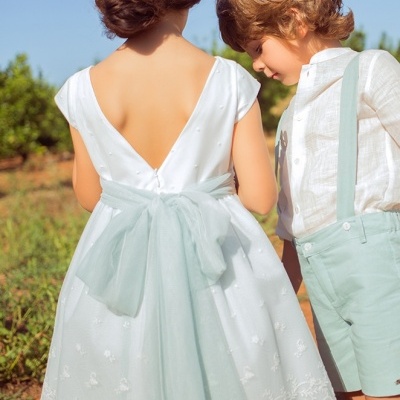 Criança vestida com vestido branco e laço azul-pálido e rapaz com camisa branca, calções azuis-pálidos e suspensórios ao ar livre