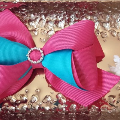 Laço de cabelo rosa e azul com anel de strass no centro sobre superfície metálica texturizada e flores ao lado