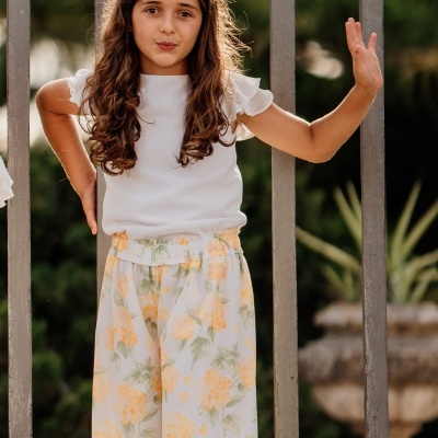 Menina com roupa branca e calças florais junto a portão metálico