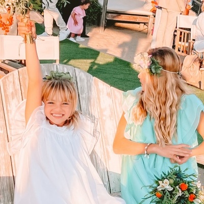 Duas meninas em cadeiras de madeira branca com vestidos brancos e verdes e coroas de flores, uma sorrindo e levantando o braço, a outra segurando bouquet colorido.