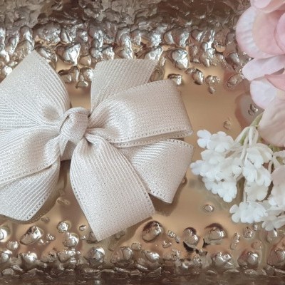 Laço branco de tecido sobre superfície dourada com textura e flores claras ao lado.