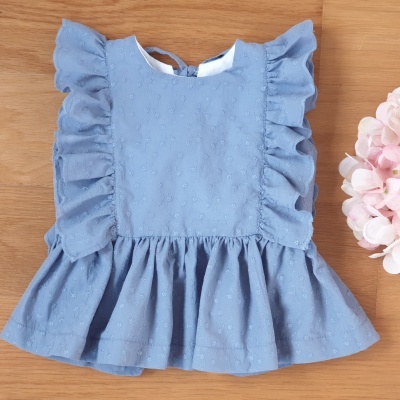 Blusa azul clara de criança com folhos e textura de pontos sobre superfície de madeira ao lado de flores cor-de-rosa