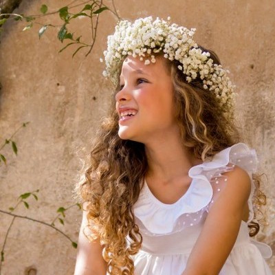 Criança com vestido branco e coroa de flores sentada junto a parede rústica