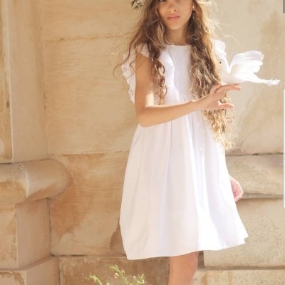 Menina com vestido branco e coroa de flores, em ambiente exterior com parede de pedra clara