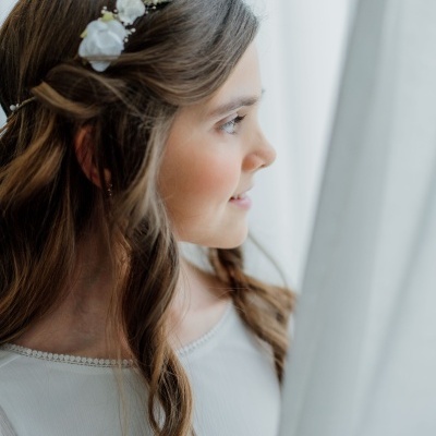 Menina com faixa de cabelo floral branca e blusa branca junto a uma cortina