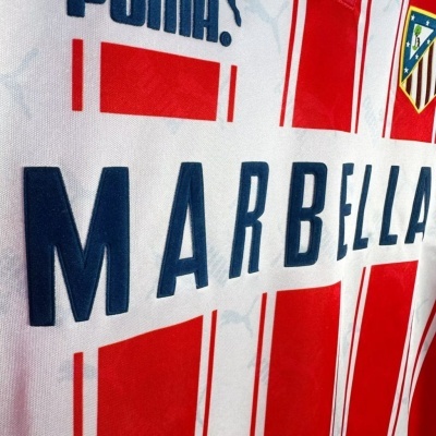 Camisola de futebol branca e vermelha com texto MARBELLA, logótipo PUMA e brasão Atlético Madrid.