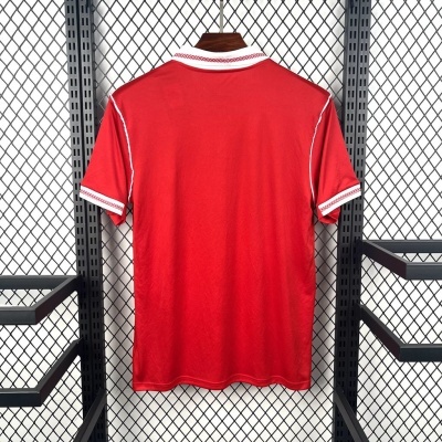 Camisola desportiva vermelha com gola e punhos brancos e padrão, pendurada contra parede metálica
