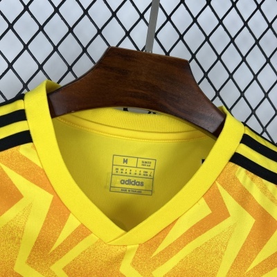 T-shirt Adidas amarela com padrão geométrico, gola em V, três riscas pretas nos ombros sobre cabide de madeira