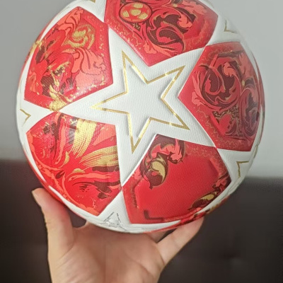 Bola de futebol com padrão vermelho, branco e dourado e estrela central segurada por mão com relógio preto