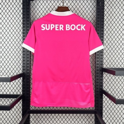Camisola rosa com texto SUPER BOCK branco, gola e mangas brancas, pendurada em suporte metálico.