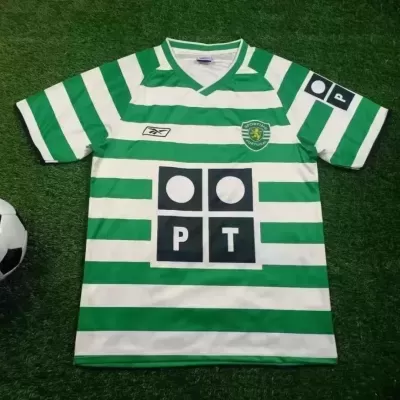 Camisola de futebol verde e branca às riscas com logótipos e bola ao lado numa superfície de relvado artificial.