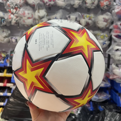 Bola de futebol branca com estrelas amarelas e vermelhas segurada por uma mão em loja com várias bolas embaladas em plástico ao fundo.