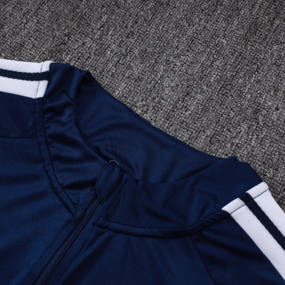 Peça de vestuário desportivo azul escuro com zipper e faixas brancas nas mangas sobre tecido cinza
