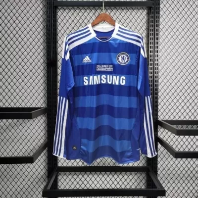 Camisola azul de futebol com logos Adidas e Chelsea, texto SAMSUNG, pendurada em estrutura metálica preta.