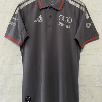 Polo cinzento escuro com logos refletivos da Audi, Revolut e Adidas, com detalhes vermelhos na gola e mangas, pendurado numa parede branca de tijolos