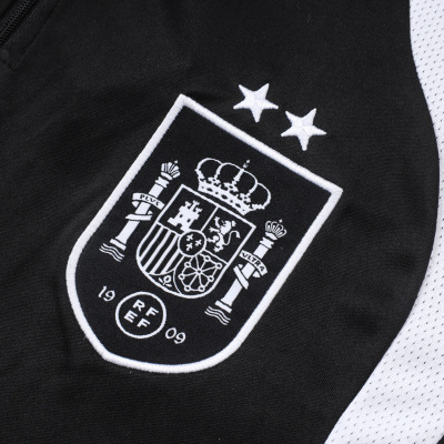 Detalhe de vestuário desportivo preto com emblema bordado branco e inserções laterais brancas perfuradas