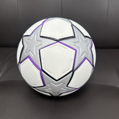 Bola de futebol branca com padrões cinza e detalhes roxos sobre sofá cinza escuro.