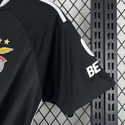 Camisola desportiva preta com detalhes brancos e emblema colorido, pendurada em fundo de grade metálica