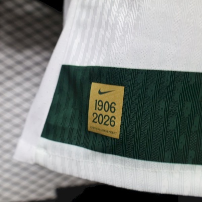 Etiqueta dourada em peça de vestuário branca com faixa verde escura e texto Nike 1906 2026