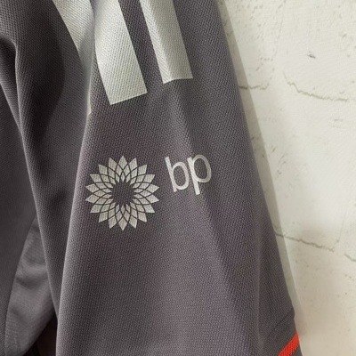 Manga de peça de roupa cinzenta com riscas brancas e logo BP, textura de malha e risca vermelha na borda.