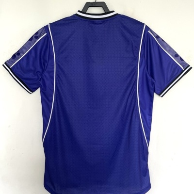 T-shirt desportiva azul com detalhes brancos, pendurada em cabide preto contra fundo branco.