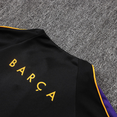 Peça de roupa preta com texto amarelo e detalhes em amarelo e roxo sobre tapete cinza.