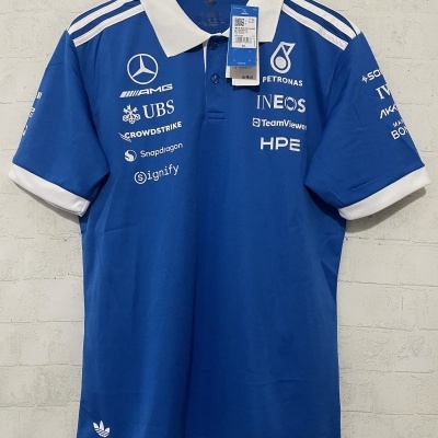 Polo azul com gola branca, múltiplos logótipos de marcas brancos e etiquetas penduradas num cabide de madeira
