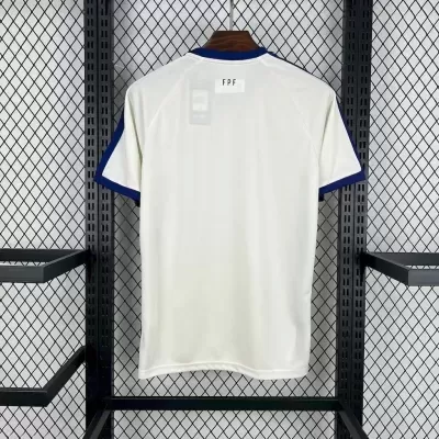 T-shirt branca com gola e mangas azuis pendurada numa estrutura metálica contra uma parede de grelha.