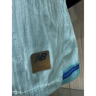Detalhe de tecido branco com padrão texturizado e etiqueta castanha NB Authentic Licensed Product