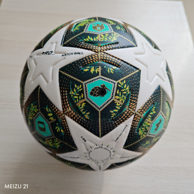 Bola de futebol branca com padrões hexagonais verde, preto, dourado e texto sobre superfície de madeira clara.