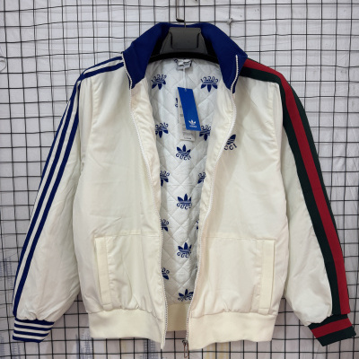 Casaco desportivo branco com padrão das marcas Adidas e Gucci no interior, riscas azuis nas mangas e riscas verdes e vermelhas noutra manga, pendurado em cabide preto contra rede branca.