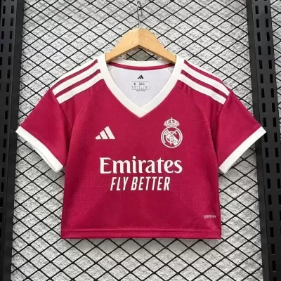 T-shirt cropped de futebol vinho com riscas brancas, logótipos Adidas e Real Madrid e texto Emirates FLY BETTER em branco, pendurada numa rede preta.