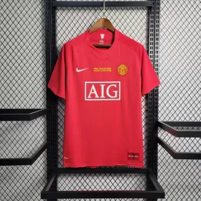 Camisola vermelha do Manchester United com logótipos e texto, pendurada em estrutura metálica preta.
