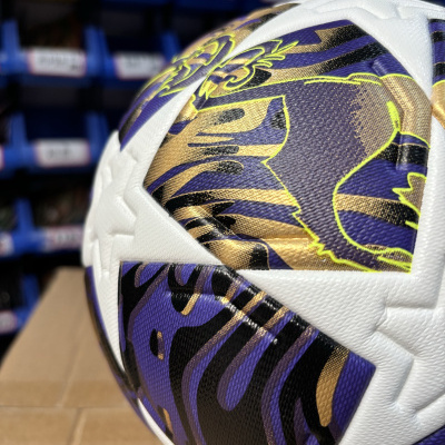 Bola de futebol branca com detalhes roxos, dourados e pretos sobre prateleiras azuis desfocadas