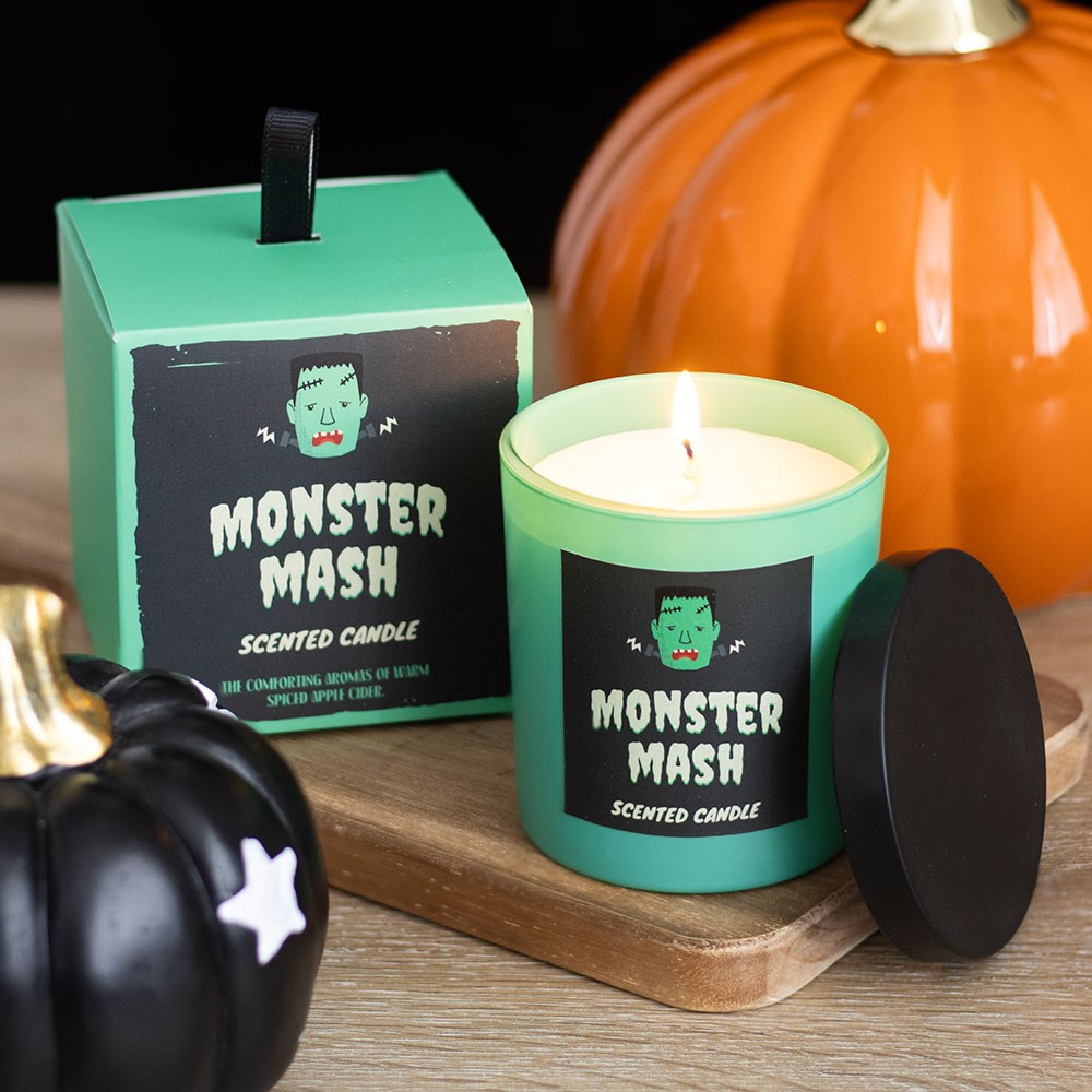 Vela aromática verde com rótulo de monstro e caixa correspondente sobre suporte de madeira com abóboras decorativas.