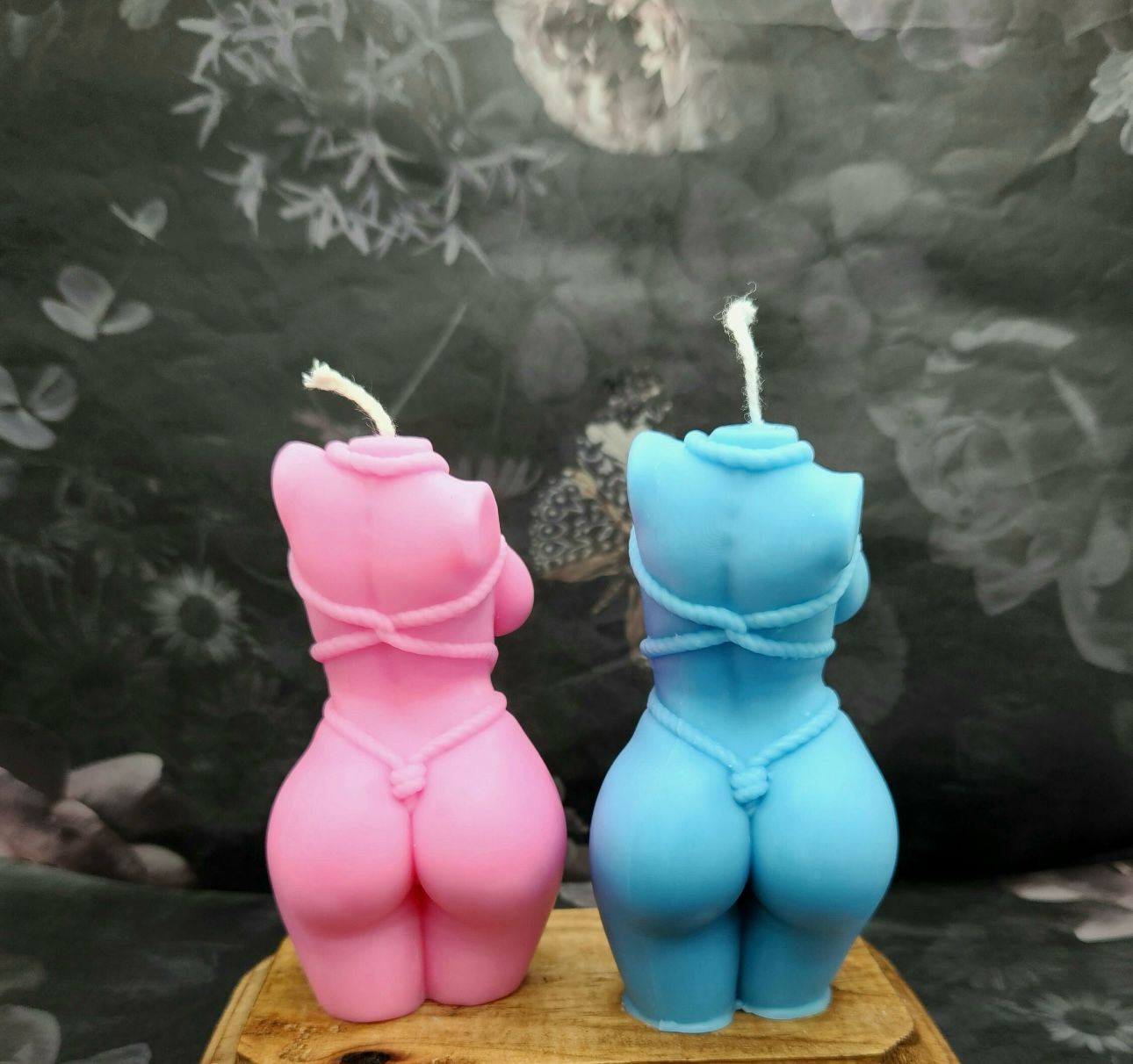 Vela Aromática Corpo Feminino Velas em forma de corpo feminino rosa e azul sobre base de madeira com fundo floral