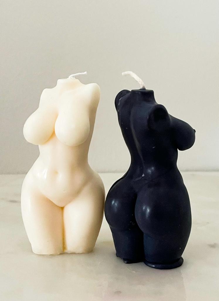 Vela Aromática Corpo Feminino Duas velas decorativas em forma de corpo feminino, uma branca e uma preta, sobre uma superfície clara.