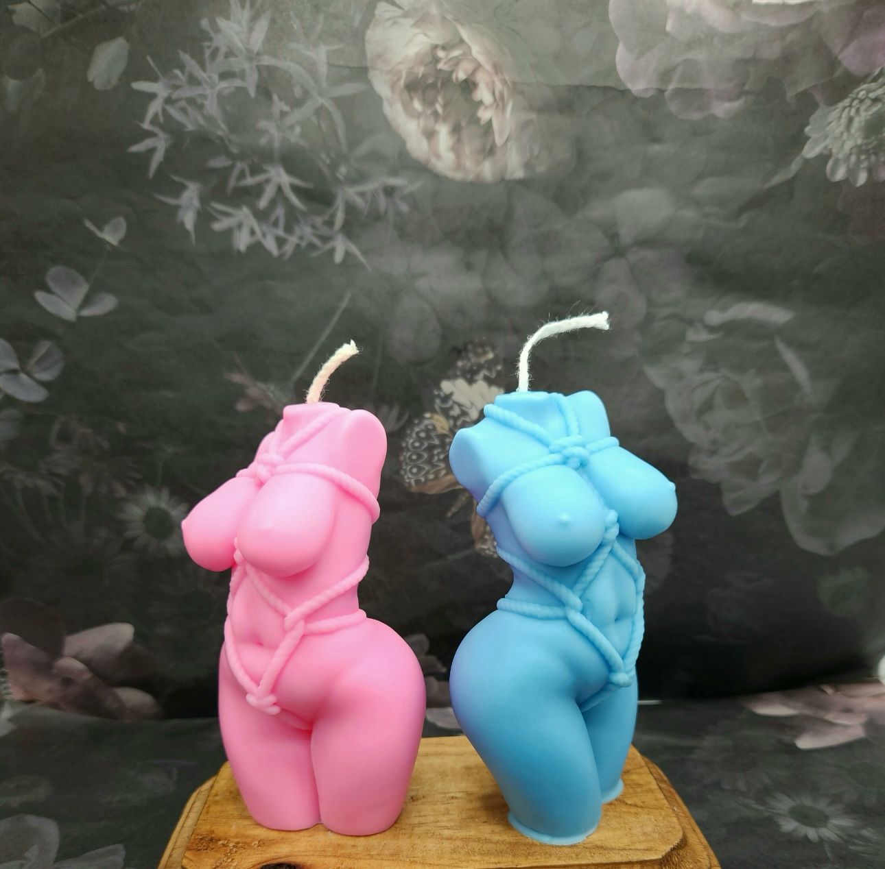 Vela Aromática Corpo Feminino Velas em forma de corpo feminino em rosa e azul com cordas, sobre base de madeira e fundo floral.