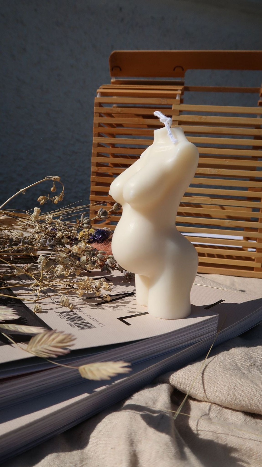 Vela Aromática Grávida Vela decorativa formato torso feminino em cera branca com flores secas e mala de madeira ao fundo