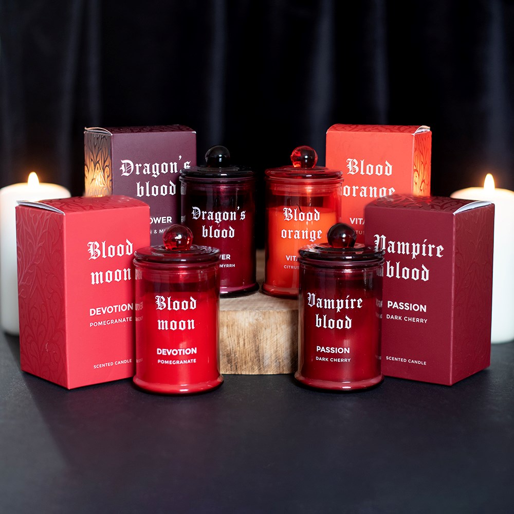 Velas perfumadas em frascos de vidro vermelho com tampas e caixas impressas com variedade de fragrâncias e nomes, dispostas sobre a mesa com fundo escuro