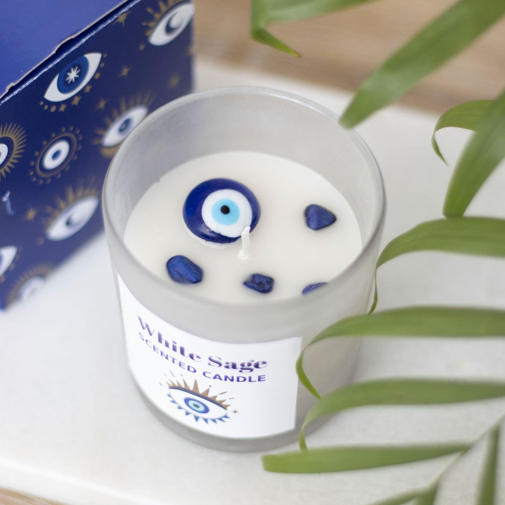 Vela perfumada White Sage com decoração de olho grego e pedras azuis, caixa azul ao fundo e folhas verdes ao lado.