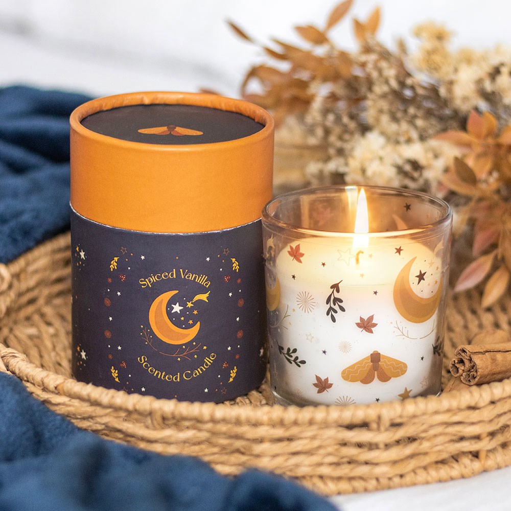 Vela aromática Spiced Vanilla com decoração de estrelas e lua, ao lado de embalagem cilíndrica, numa bandeja de palha com fundo azul e raminhos secos.