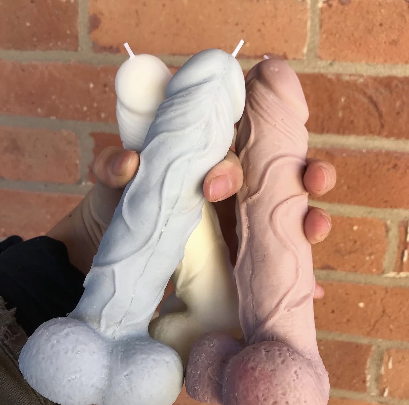 Vela Aromática Pénis Velas em formato fálico em azul claro, branco e rosa sendo seguradas por uma mão com parede de tijolos ao fundo