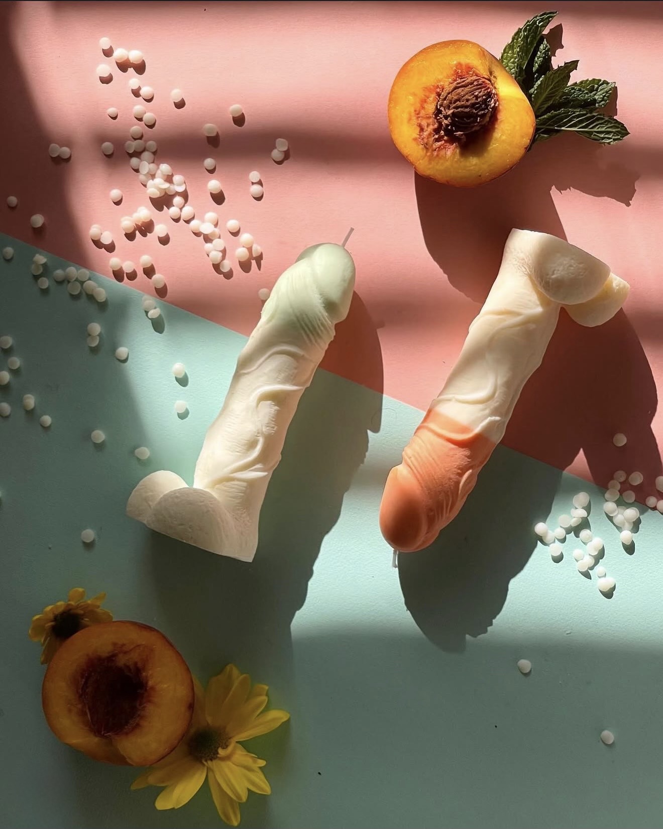 Vela Aromática Pénis Duas velas em forma de pénis com pêssegos, flores amarelas e bolinhas brancas sobre fundo rosa e azul